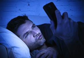 A man lying in bed looks at his smartphone screen with a thoughtful expression, illuminated by its blue light in a dark room, possibly searching for SEO keywords.