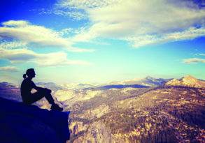 A person sitting on a rocky ledge overlooking the vast scheme of a mountainous landscape under a clear, blue sky.