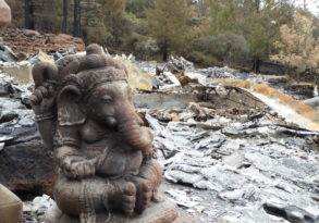 A statue of Ganesha amid the debris of a burned-down area, with charred remains and scattered ashes echoing its last words in the background.