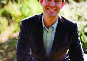 A smiling man in a blazer and shirt sitting outdoors with sunlight filtering through trees in the background, similar to an image you might find in an Ocean Robbins book.