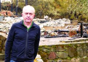 Man standing in front of a ruined retreat with stone walls, wearing a black jacket and looking directly at the camera.