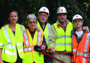 Five construction workers in safety vests and helmets stand together, holding tools, against a backdrop of green foliage that paradoxically thrives amidst urban development.