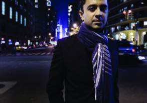 Man in a dark jacket and scarf standing on a city street at night, with illuminated buildings and streets in the background, exuding an "Iyer on Fire" vibe.