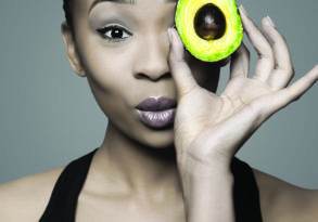 A Black girl holds a halved avocado over one eye, gazing into the camera.