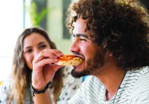 A young man with curly hair enjoying a slice of pizza, with a young woman smiling in the background. They clearly have the munchies.