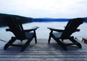 Two adirondack chairs facing a tranquil lake, set on a wooden dock with a forested hill in the background, offering perfect relaxation.