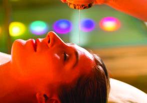 A woman lies on her back receiving an essential oil treatment on her forehead, with colorful blurred lights in the background, setting a course for serenity.