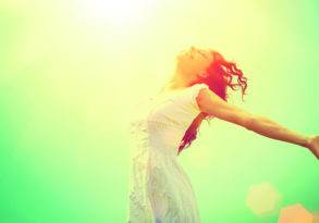 A woman in a white dress joyfully stretching her arms with sunlight bathing her, against a bright green background, embodying the essence of setting healthy boundaries.