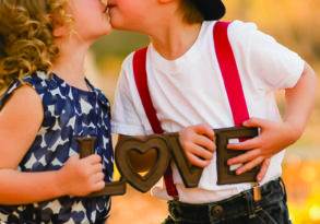 Two children in an outdoor setting, a boy wearing a hat and suspenders sharing his first kiss with a girl in a blue dress, both holding heart-shaped objects.