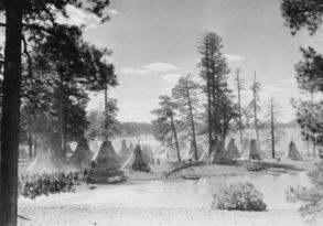 Black and white historical photo of a native American encampment with teepees near a body of water, surrounded by forest and a gathering of people sharing last words.