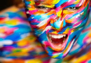 A person with vibrant, multicolored face and body paint, smiling with visible enthusiasm.