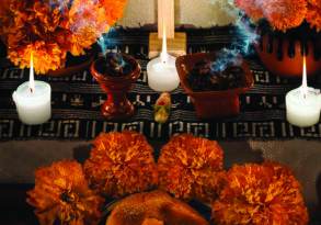 A traditional day of the dead altar remembering ancestors, featuring lit candles, orange marigolds, a wooden cross, smoking copal incense burners, bread, and a decorative skull.