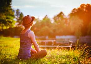 A person in a hat and tank top reconnecting with nature while sitting on grass in a park during sunset, facing away from the camera.