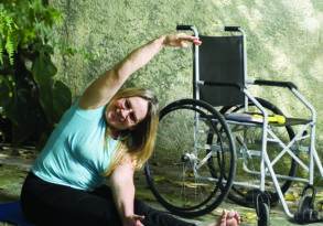 Woman in exercise attire practicing accessible yoga on a blue mat next to her wheelchair, in a garden with a stone wall and fallen leaves.