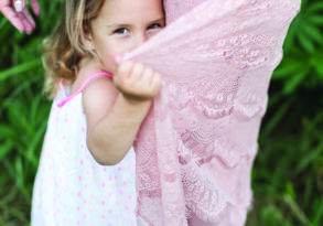 A young girl in a polka dot dress holding hands with an adult, partially hidden behind the adult's pink lace dress, communicating through a secret language as they look at the camera.