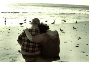 Two people sitting closely on a beach, embracing as they share last words while watching seagulls on the sand, with the ocean in the background.