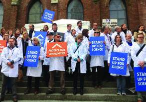 Group of scientists in white lab coats holding protest signs with messages like "protect our communities" and "stand up for scientific integrity" on steps of a brick building.